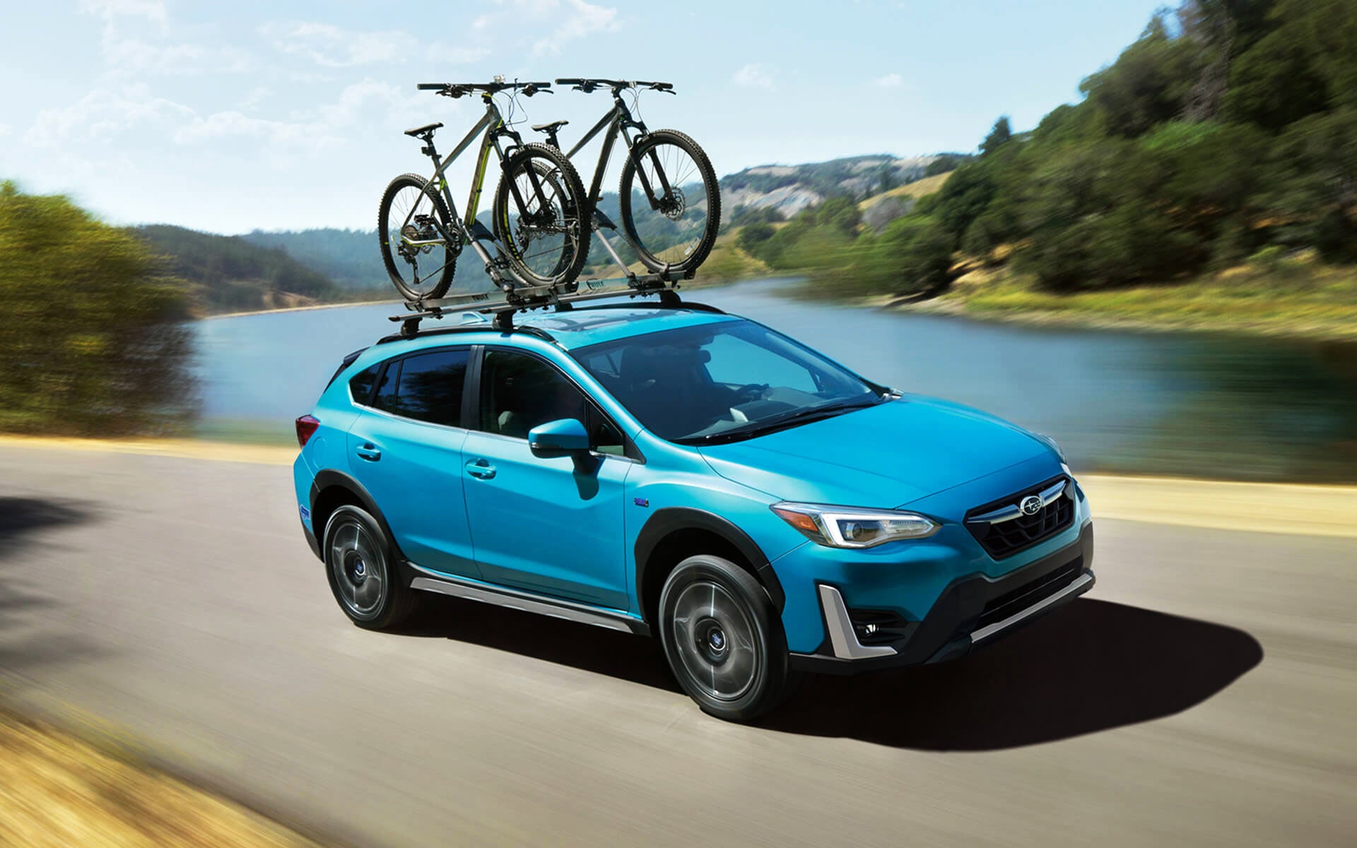 A blue Crosstrek Hybrid with two bicycles on its roof rack driving beside a river | LaFontaine Subaru in Commerce Township MI